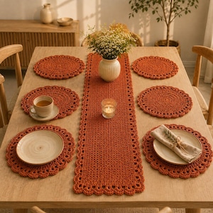 May include: A set of burnt orange crochet placemats and a matching rectangular table runner with a scalloped edge. The table setting includes ceramic plates, a tea cup on a saucer, a linen napkin tied with twine, and a white vase with flowers.