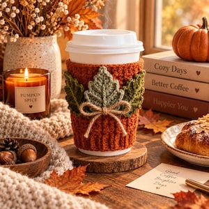 Peut inclure: Un gobelet à café blanc jetable doté d'une housse en crochet orange brûlé texturée, ornée de trois feuilles crochetées vertes et d'un nœud en ficelle de jute. Le gobelet repose sur un sous-verre en bois rond, entouré de décorations automnales.