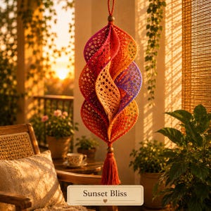 Pode incluir: Um ornamento espiral suspenso com um gradiente de padrões de crochê laranja, vermelho e roxo com recortes circulares intrincados. A peça é suspensa por um cordão com uma conta de madeira e finalizada com uma borla laranja a condizer.
