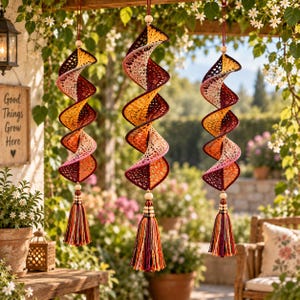 May include: Three hanging spiral ornaments crafted from crocheted yarn in a gradient of deep burgundy, orange, and cream. Each piece features a central wooden bead at the top and bottom, finished with a multi-colored yarn tassel at the base.