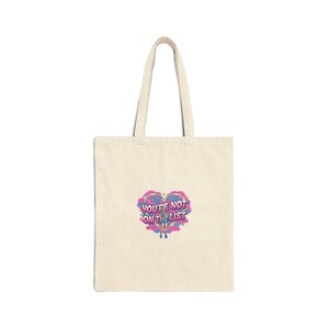 Pode incluir: Saco tote creme com um gráfico em forma de coração que diz "YOU'RE NOT ON THE LIST" em uma fonte estilizada e colorida. O saco tem alças longas e é feito de um tecido resistente.