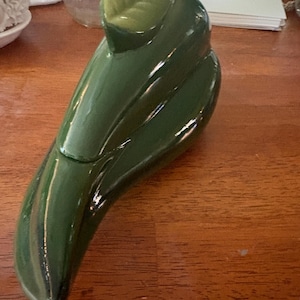 Pode incluir: Um recipiente de cerâmica verde-escuro brilhante com tampa, em forma de pimenta longa e curva. A tampa apresenta um detalhe de folha esculpida em verde mais claro no topo. O objeto tem um acabamento liso, reflexivo e forma alongada.
