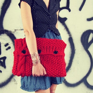 May include: A red knitted clutch bag with a black button closure. The bag is held by a person wearing a black and white polka dot top and a denim skirt.