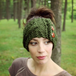 May include: A green knitted headband with brown button accents. The headband is worn by a person with brown hair and brown eyes.