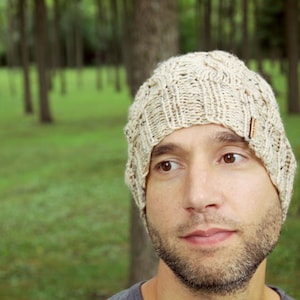 May include: A close-up of a person wearing a beige knitted beanie with a cable knit pattern. The beanie has a brown leather tag on the side.