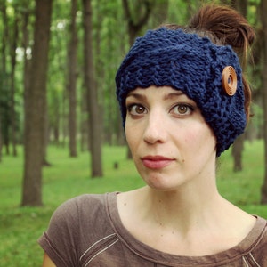 May include: A woman wearing a blue knitted headband with a brown button on the side. The headband is covering her ears. She is looking at the camera.
