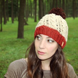 Op de afbeelding: Een vrouw draagt een witte en rode gebreide muts met een bruine pompon. De muts heeft een kabelbreipatroon en een wit label.