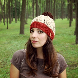 Op de afbeelding: Een vrouw draagt een gebreide muts met een wit en rood kleurenschema. De muts heeft een pompon bovenop en een bruin lederen label.