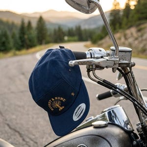 Peut inclure: Casquette de baseball bleu marine avec le texte "Russet & Rune" et un motif d'arbre doré. La casquette est accrochée au guidon d'une moto, avec une route et des montagnes en arrière-plan.