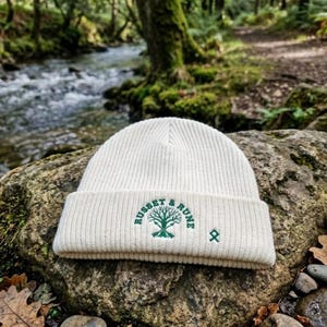 May include: Off-white ribbed knit beanie hat featuring the words "RUSSET & RUNE" and a green tree graphic. A small green symbol is to the right of the text. The hat is shown on a rock in a natural outdoor setting with a stream in the background.