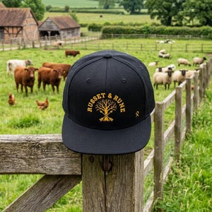 May include: Black baseball cap with gold "RUSSET & RUNE" text and a tree logo. The cap is on a wooden fence, with a rural scene of fields, cows, and sheep in the background. The cap is a classic style.