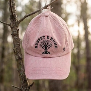 Peut inclure: Casquette de baseball rose poudré avec le texte "RUSSET & RUNE" et un motif d'arbre. La casquette est suspendue à une branche d'arbre. L'arrière-plan est flou, avec des arbres et la lumière du soleil.