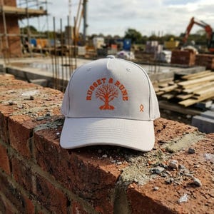 Puede incluir: Gorra de béisbol gris claro con el texto naranja "RUSSET & RUNE" y un diseño de árbol. La gorra está colocada sobre un muro de ladrillos rojos, con maquinaria de construcción al fondo.