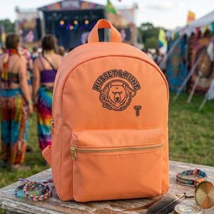 May include: A solid peach-coloured backpack featuring a black graphic print of a bear head inside a circular frame with the text "RUSSET & RUNE" above it and a small symbol below. The backpack has a front zippered pocket and gold-tone hardware.