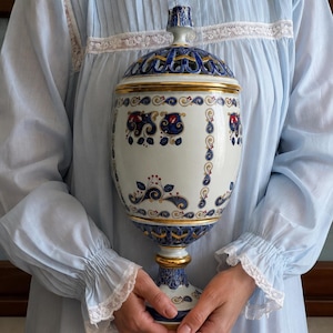 Może przedstawiać: Ozdobna ceramiczna urna z pokrywką, posiadająca białą podstawę z misternymi, malowanymi wzorami kwiatowymi i zawijasami w kolorach niebieskim, czerwonym i złotym. Przedmiot ma stopkę oraz ażurowy wzór kratki na pokrywce i dolnej części.