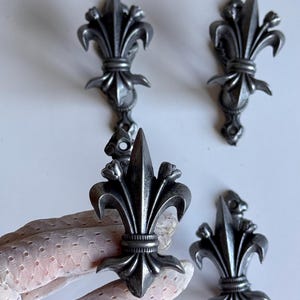 Op de afbeelding: Een set van vier zilverkleurige metalen ornamenten in de vorm van een fleur-de-lis. Elk stuk heeft een puntige centrale piek geflankeerd door twee gebogen bloemblaadjes, met een horizontale band aan de basis en een klein rond montagegat.