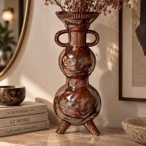 Può includere: Un alto vaso in ceramica color marrone metallizzato con una silhouette impilata e bulbosa, due manici laterali e tre gambe corte. La superficie presenta una texture martellata e riflettente. Fogliame marrone essiccato è disposto all'interno del vaso.