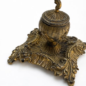 Pode incluir: Um tinteiro de metal dourado de estilo antigo com uma base quadrada ornamentada com volutas intrincadas e detalhes em forma de folha. O reservatório central é rematado por uma tampa abobadada a condizer com uma pequena pega curva.