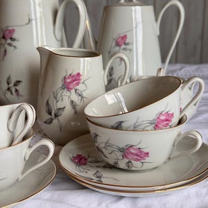 May include: A porcelain tea set featuring off-white pieces decorated with pink rosebuds and grey stems. The set includes two tall teapots with lids, a creamer, and several teacups with matching saucers, all finished with thin gold trim.