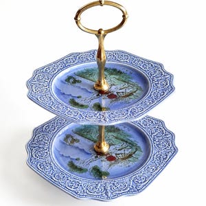 Puede incluir: Un soporte de dos niveles con dos platos octogonales de cerámica azul, bordes en relieve detallados y una ilustración de paisaje central. Los niveles están unidos por un asa central de metal dorado con un lazo ovalado.