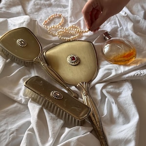 Könnte beinhalten: Ein antikes Schminkset mit Handspiegel, Bürste und kleinem Parfümflakon. Das Set ist goldfarben mit dekorativen Details und einem roten Schmuckstein. Eine Perlenkette und ein weißes Tuch vervollständigen die Szene.