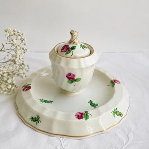 Peut inclure: Un sucrier en porcelaine blanche avec couvercle et sa soucoupe assortie, orné d'un motif peint à la main de roses roses et de feuilles vertes. Les pièces présentent un design cannelé à bords festonnés avec un liseré doré et un bouton torsadé.