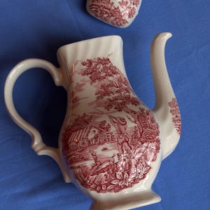 Può includere: Teiera in ceramica bianca con un design a scena pastorale rossa, tra cui un cacciatore, un cervo e una capanna. La teiera ha un manico e un beccuccio curvi, con un coperchio abbinato. Il design è un classico motivo toile.