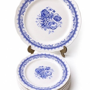 Op de afbeelding: Een set witte keramische borden met een blauw bloemmotief in het midden en een blauwe rasterrand langs de geschulpte randen. De borden zijn gestapeld, waarbij één bord rechtop op een metalen standaard wordt getoond voor het ontwerp.