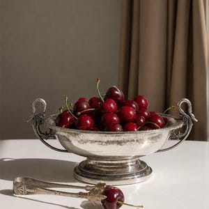 Peut inclure: Un bol sur pied en métal argenté avec des anses ornées en forme de cygne, rempli d'un tas de cerises fraîches rouge foncé. Une paire de pinces de service assorties en métal argenté repose devant le bol, tenant une cerise rouge foncé.