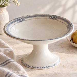 Pode incluir: Uma taça de cerâmica cor de creme sobre pedestal com uma base robusta e um recipiente largo e raso. A borda e a base são decoradas com um delicado padrão azul composto por pequenos motivos florais e bordas lineares detalhadas.