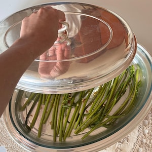 Könnte beinhalten: Eine Servierschale aus klarem Glas mit silbernem Rand, gefüllt mit grünem Spargel. Die Schale ist mit einem Deckel aus klarem Glas mit einem silbernen Griff bedeckt. Die Schale steht auf einer weißen Oberfläche.