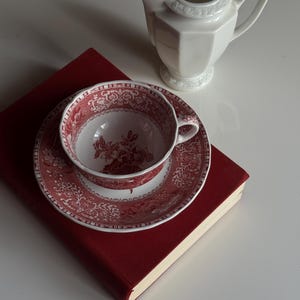 May include: A white creamer and a teacup and saucer set with a red floral pattern sit atop a red book. The teacup and saucer have a detailed design. The creamer has a geometric shape.