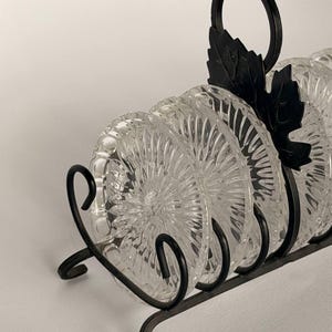 May include: A set of clear glass coasters with a sunburst pattern, held in a black metal rack. The rack features a decorative handle shaped like a leaf and curved metal supports that hold the circular glass coasters in an upright position.
