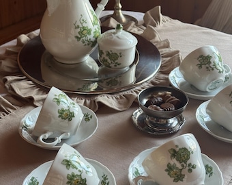 May include: A white porcelain tea set with a green floral pattern. The set includes a teapot, sugar bowl, and several teacups with saucers. The teacups are arranged on a table with a silver tray and a small bowl of chocolates.