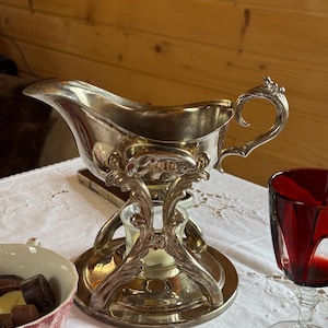 Puede incluir: Salsera plateada con detalles ornamentados, sobre un soporte a juego. La salsera tiene un pico curvo y un asa decorativa. El soporte presenta una base circular y volutas intrincadas. También se ven una copa de vino tinto y un cuenco de bombones.