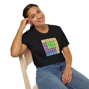 May include: Black t-shirt with a colorful word puzzle design. The puzzle spells out "TREASURE MAP" in a grid of pastel-colored squares. The shirt is worn by a person sitting in a wooden chair.