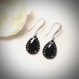 May include: A pair of silver-toned drop earrings with black teardrop-shaped stones. The earrings have a small, clear stone accent on the hook. The black stones are surrounded by a row of smaller black stones.