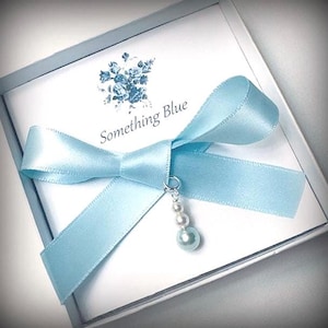 May include: A light blue gift box with a satin ribbon tied in a bow. The box contains a card with the words "Something Blue" and a floral design. A charm with pearls hangs from the bow.