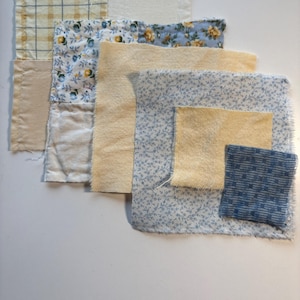 May include: A collection of fabric swatches in various patterns and colors. The swatches include a yellow plaid, floral prints in blue and yellow, and solid beige and blue fabrics. The arrangement suggests a quilting or textile project.