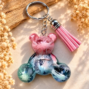 Op de afbeelding: Een hars sleutelhanger in de vorm van een teddybeer met een wervelende mix van roze, blauwe en groene kleuren met een doorschijnende afwerking. De beer is bevestigd aan een zilveren metalen ring en een korte, lichtroze kunstsuède kwastje.
