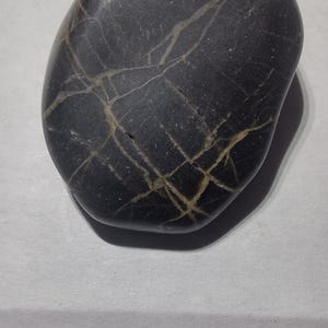 May include: A smooth, dark gray stone with a network of light-colored veins. The oval-shaped rock has a polished surface and a small hole near the top. The stone is set against a light gray background.