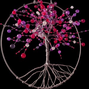 Op de afbeelding: Handgemaakte draadboomsculptuur met een zilveren frame en een zwarte achtergrond. De boom heeft een gedraaide draadstam en wortels, met takken versierd met rode, roze, paarse en heldere kralen. De boom wordt opgehangen door een zilveren ketting.