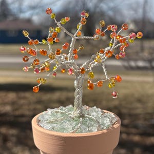 Puede incluir: Una escultura de árbol de alambre hecha a mano con cuentas de vidrio de colores que representan hojas y bayas. El árbol está plantado en una pequeña maceta de terracota llena de guijarros de vidrio transparente. Las ramas del árbol están adornadas con cuentas naranjas, amarillas y rosas.