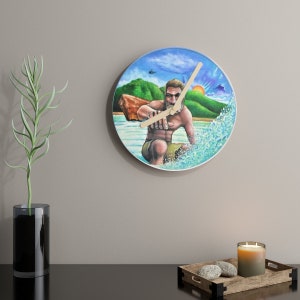Puede incluir: Un reloj redondo con una imagen pintada de una persona surfeando en una ola. El fondo es un cielo azul con nubes y una isla verde. Las manecillas del reloj son de madera.