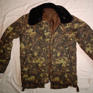 May include: A camouflage print jacket featuring a black faux fur collar and a brown interior lining. The jacket includes a front zipper closure, two slanted chest pockets with zippers, and two lower pockets with vertical zipper openings.