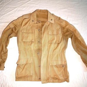 May include: Tan-colored utility jacket with four front pockets and button closures. The jacket has long sleeves and a collared neckline. The fabric appears to be a soft, worn material. The jacket is laid flat on a white surface.