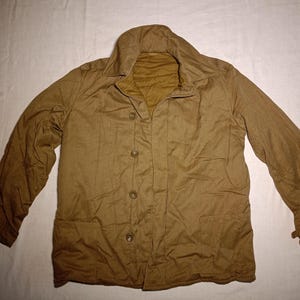May include: A tan-colored jacket featuring a wide collar, long sleeves, and a front button closure with four visible buttons. The garment includes two large patch pockets at the lower front and is constructed from a solid, matte fabric.