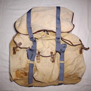 Puede incluir: Una mochila de lona beige con correas de nailon azul, ribetes de cuero marrón y hebillas de metal. La bolsa tiene una solapa superior grande, un cierre de cordón y dos bolsillos delanteros con correas de cuero, mostrando signos de desgaste y manchas.
