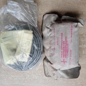 Op de afbeelding: Twee medische kits. Eén zit in een doorzichtige plastic zak, met een zwart, cirkelvormig object en verschillende gevouwen, crèmekleurige items. De andere is een bruin, rechthoekig pakket met rode tekst en een rood kruis.