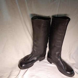 May include: A pair of tall, black leather boots featuring a textured finish, rounded toes, and a structured shaft that reaches mid-calf. The boots have a smooth, dark appearance with visible stitching details along the foot and a sturdy sole.